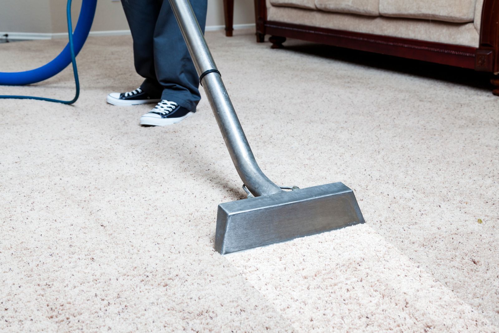 A person in blue work clothes and gloves mops a wooden floor in front of a grey sofa.