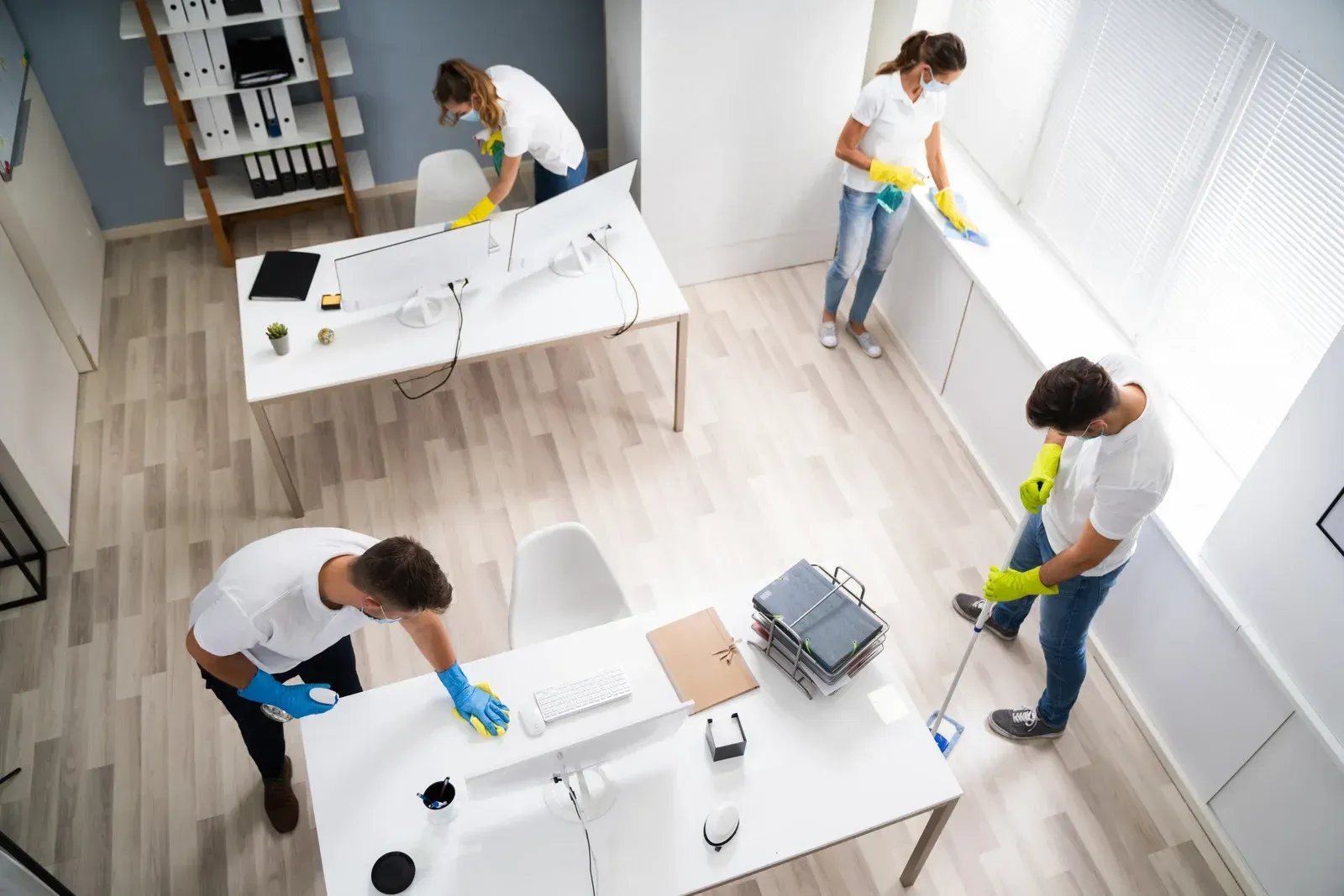 Four cleaners wearing uniforms and yellow gloves work to disinfect and clean a modern office space.
