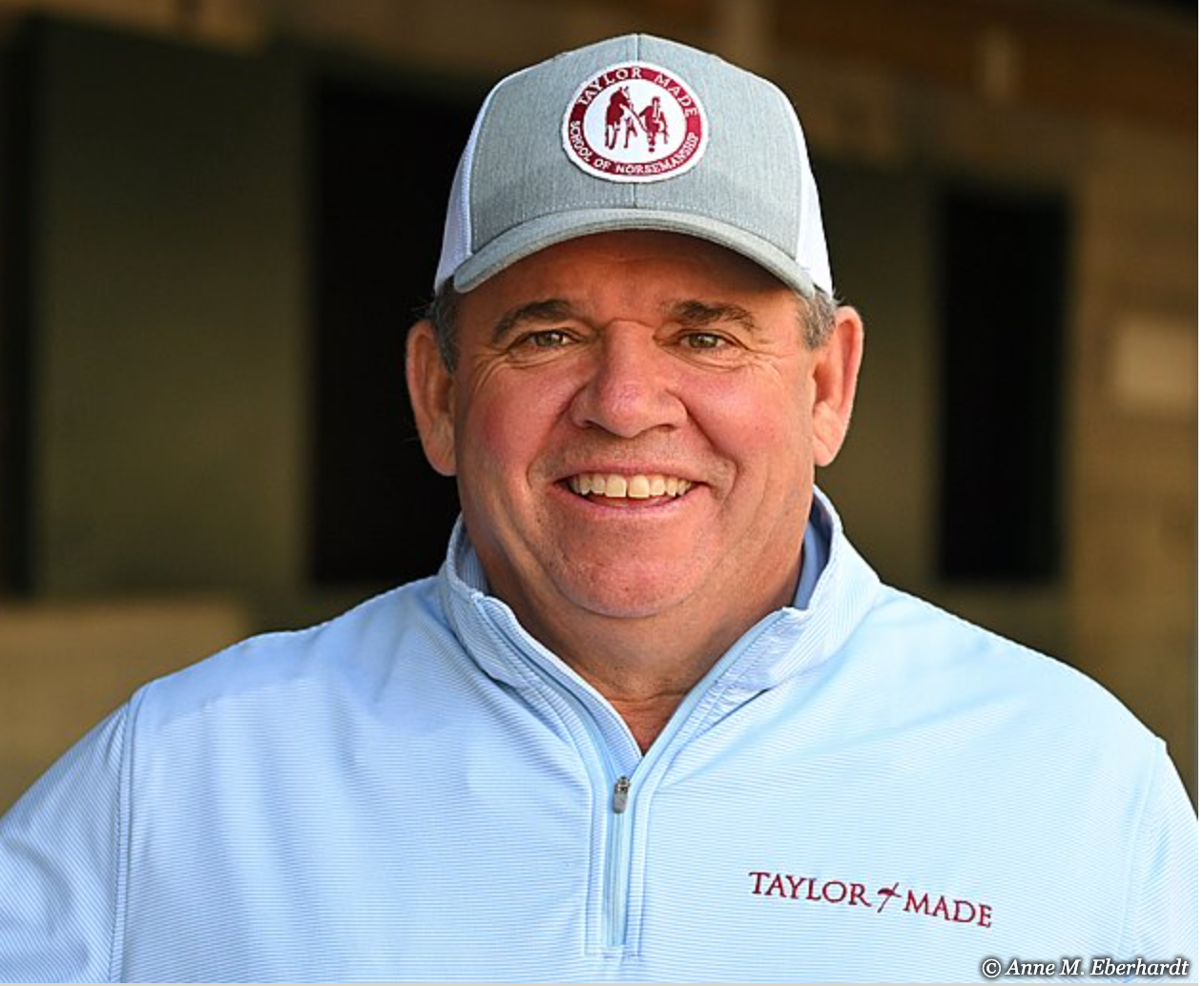 Smiling man in a blue Taylor Made jacket and hat with a logo, standing outdoors.