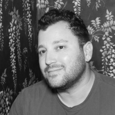 Man with dark hair and stubble smiles, poses near floral wallpaper.