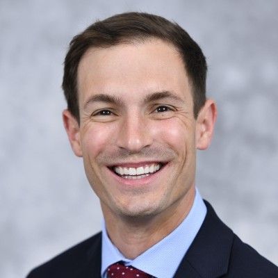 Man with brown hair and a smile, wearing a suit and tie, looking at the viewer.