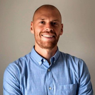 Bald man with a red beard smiles. He wears a blue button-down shirt, standing against a grey wall.
