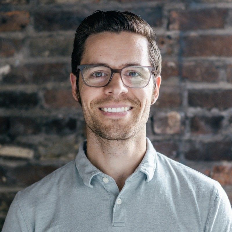 Man with glasses smiling in front of a brick wall, wearing a light blue shirt.