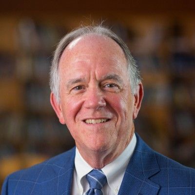 Smiling older man in a blue suit and tie, against a blurred background of books.
