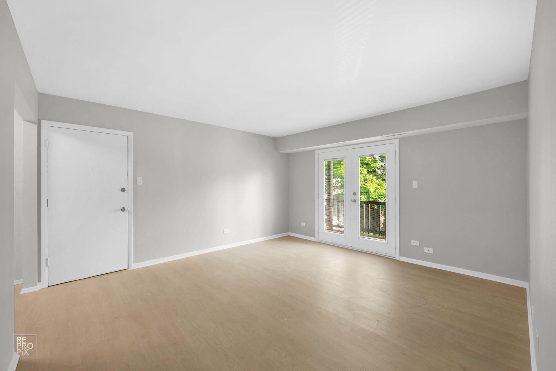 Empty, gray-walled room with light-colored floor, white door, and French doors leading to a balcony.
