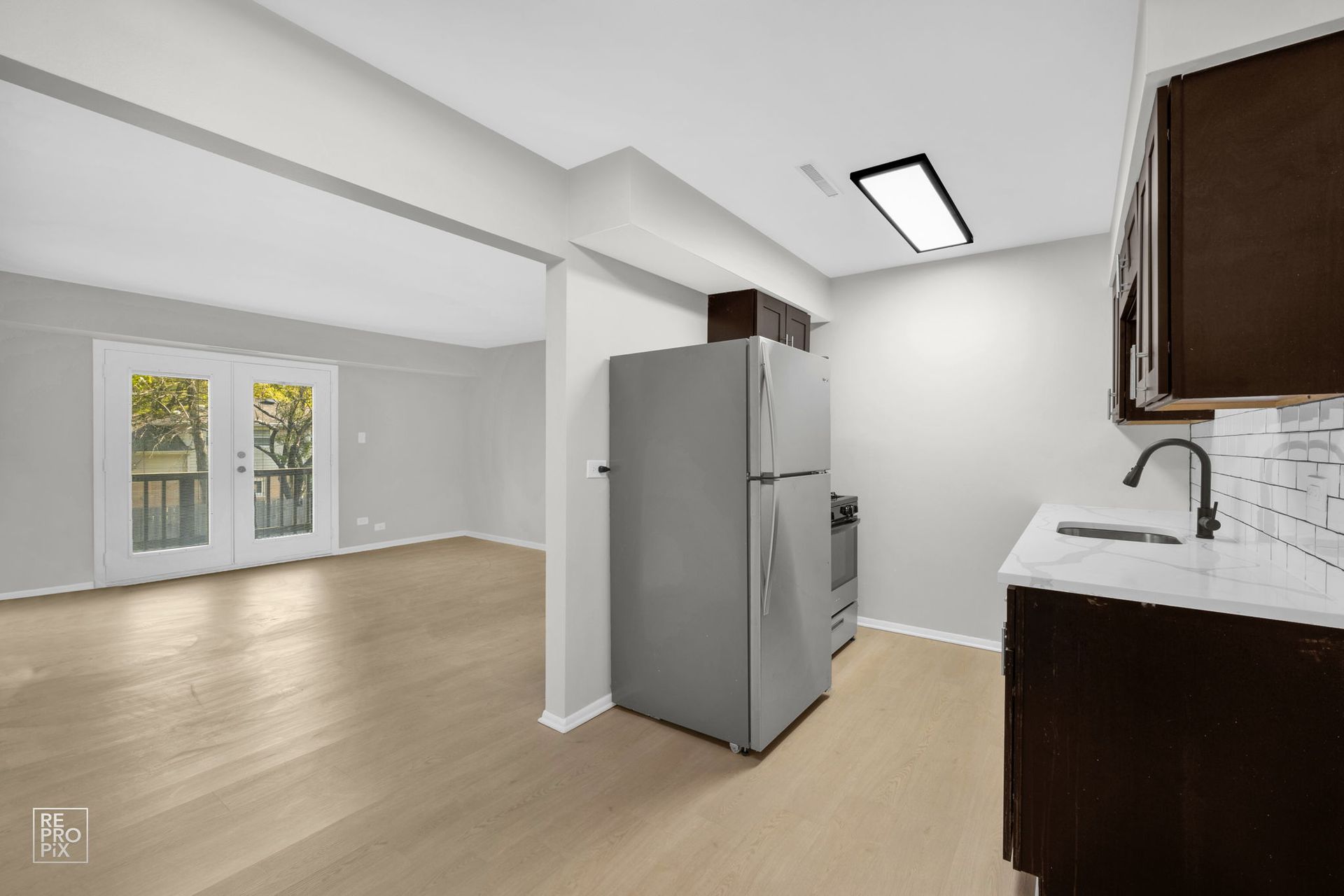 Kitchen with dark cabinets, stainless steel appliances, and a view of the living room with wood floors and a door.