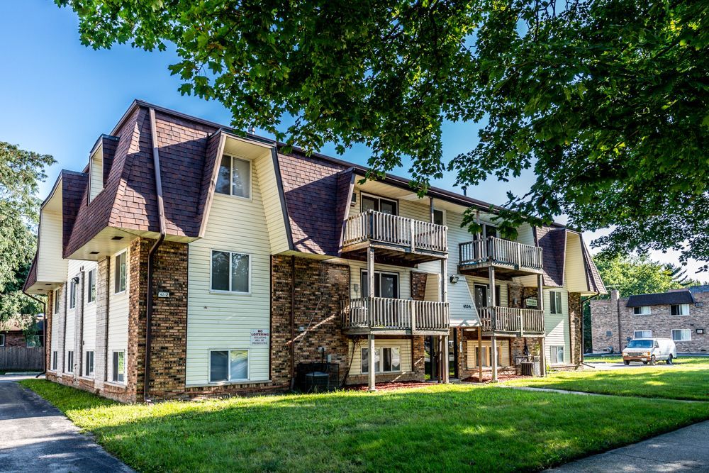Apartment building with white siding, brick, and brown roof on a sunny day, with balconies and green lawn.
