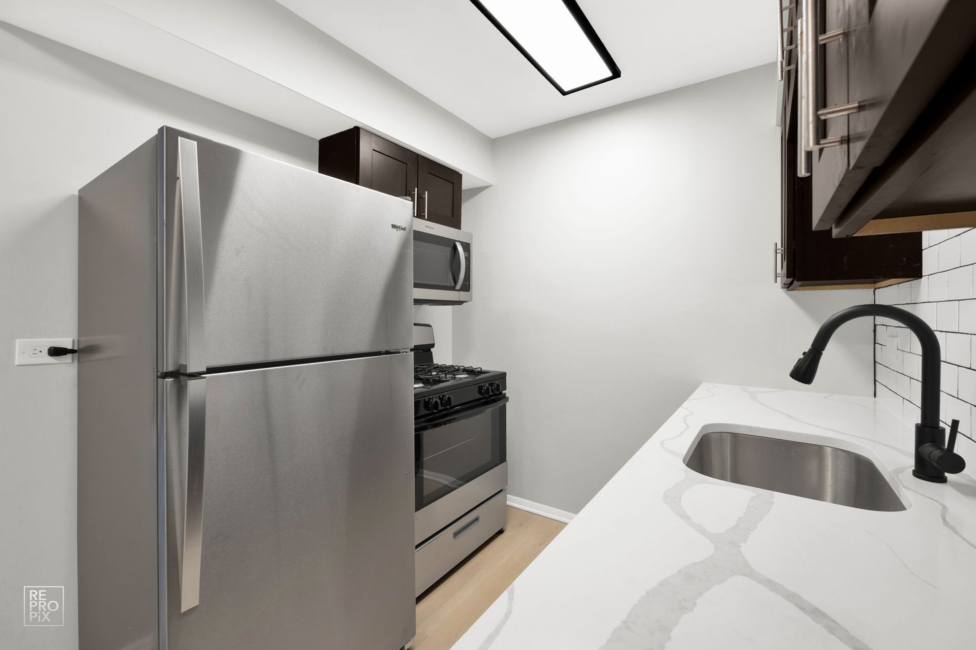 A small, modern kitchen with stainless steel appliances, white countertops, and dark cabinets.