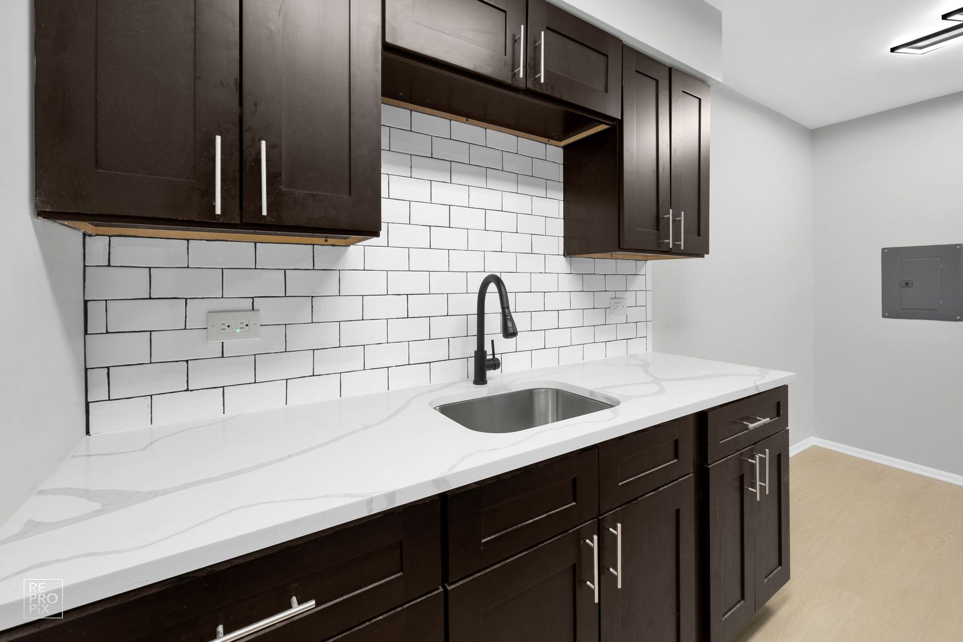 Dark brown cabinets with a white countertop and sink; white brick backsplash; black faucet.