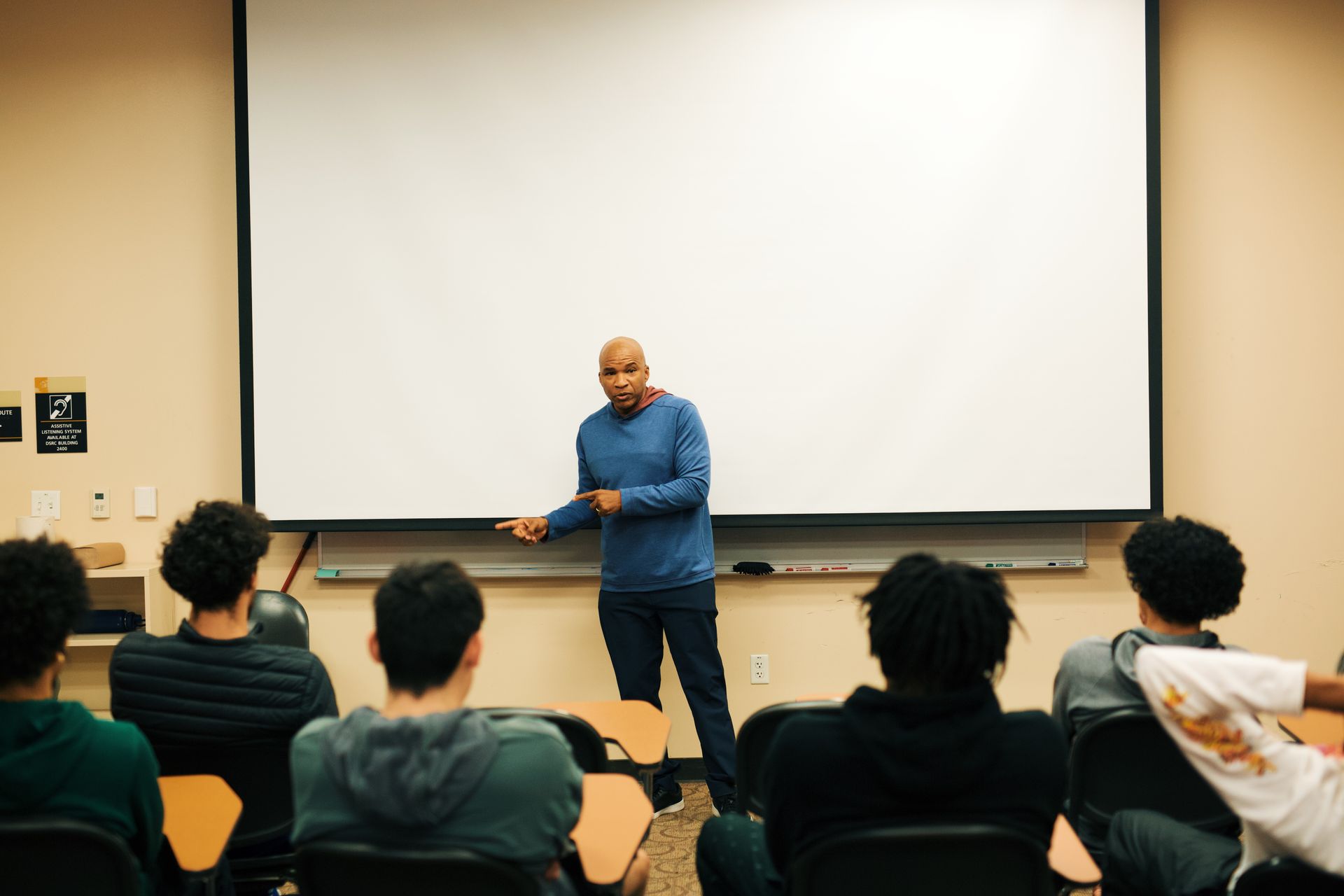 Man giving a presentation to a group of students in a classroom, pointing at the screen.