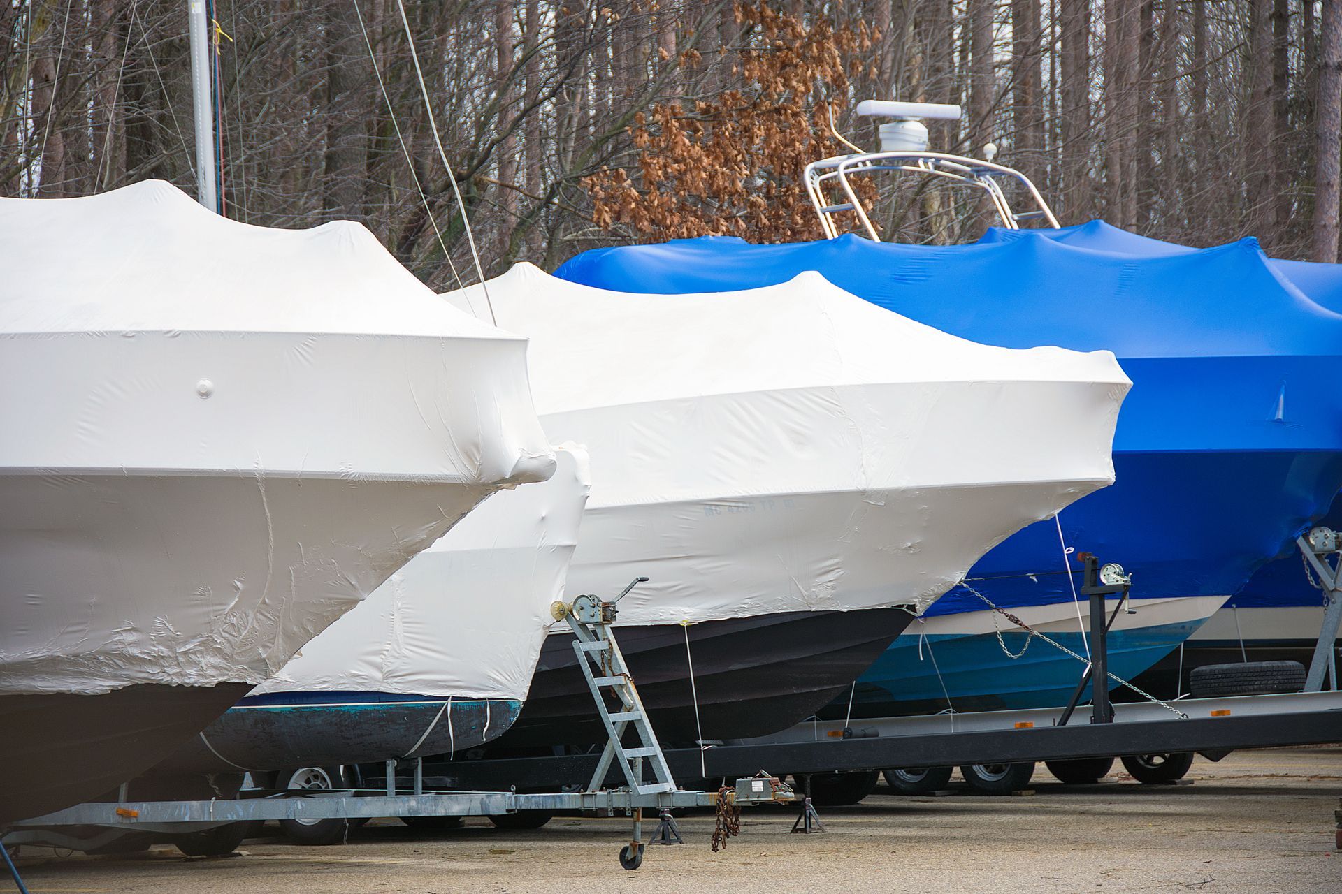 Boats covered in white and blue tarps parked on trailers in a lot.