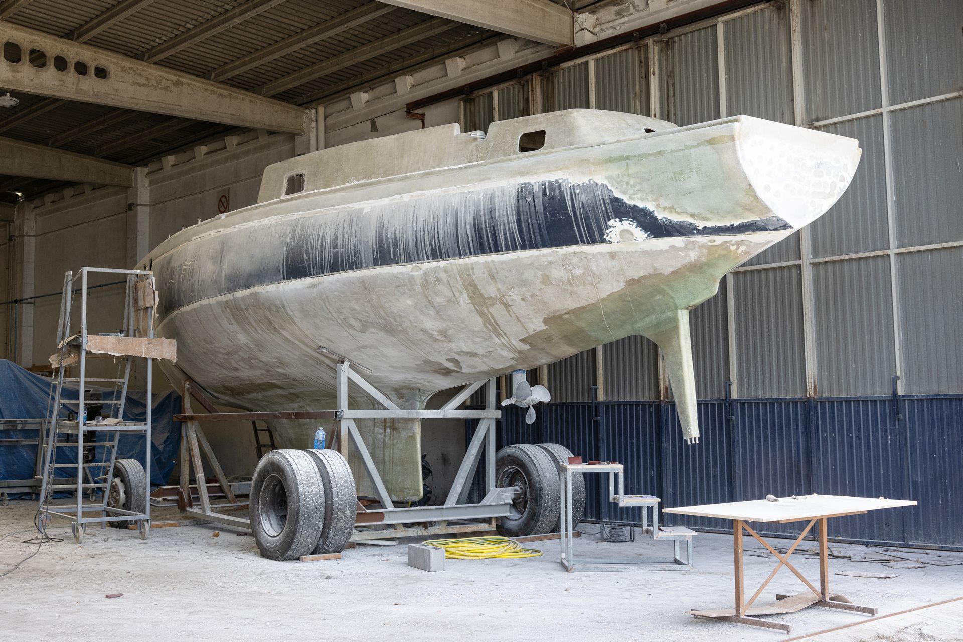 Sailboat hull on a wheeled cradle inside a workshop; shows repair work with patches.
