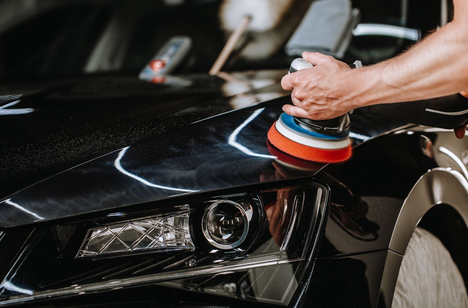 Person polishing a black car with a rotary buffer, indoors.