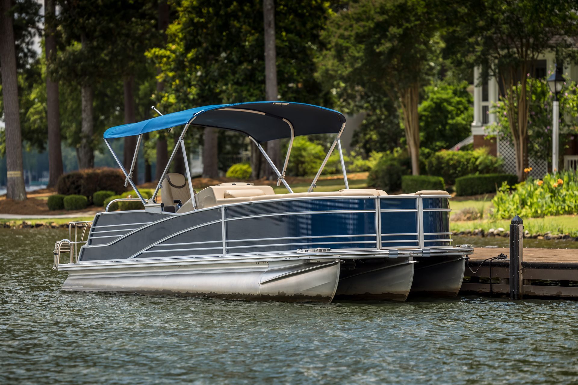 Blue pontoon boat docked on a lake, with a blue canopy. Trees and a house in the background.