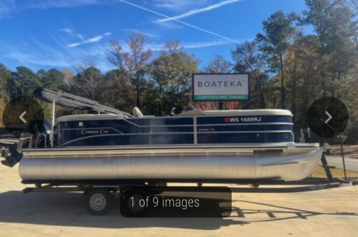 A blue and silver pontoon boat on a trailer, parked outside a Boateka sign on a sunny day.