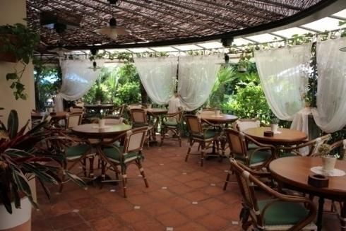 Caffè all'aperto con tavoli e sedie in legno, pavimento in terracotta e tende bianche sotto una tettoia ombreggiata.