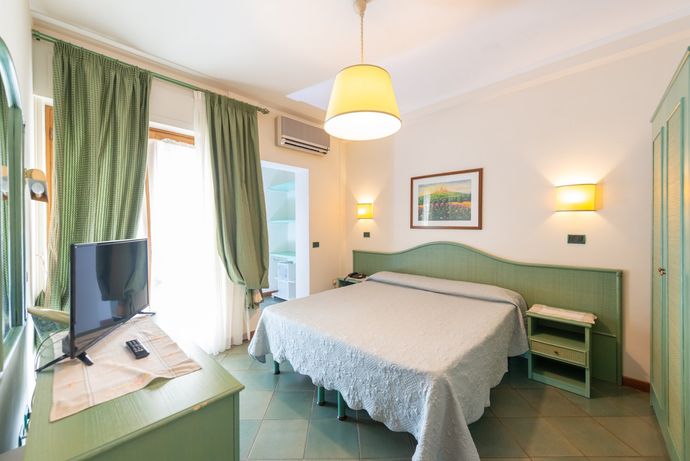 Luminosa camera d'albergo con letto, tende verdi, TV, scrivania e lampade a parete.