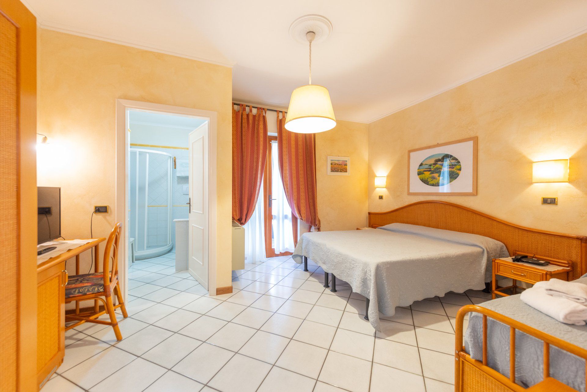 Luminosa camera d'albergo con due letti singoli, pavimento piastrellato, scrivania, lampade e porta del bagno aperta.