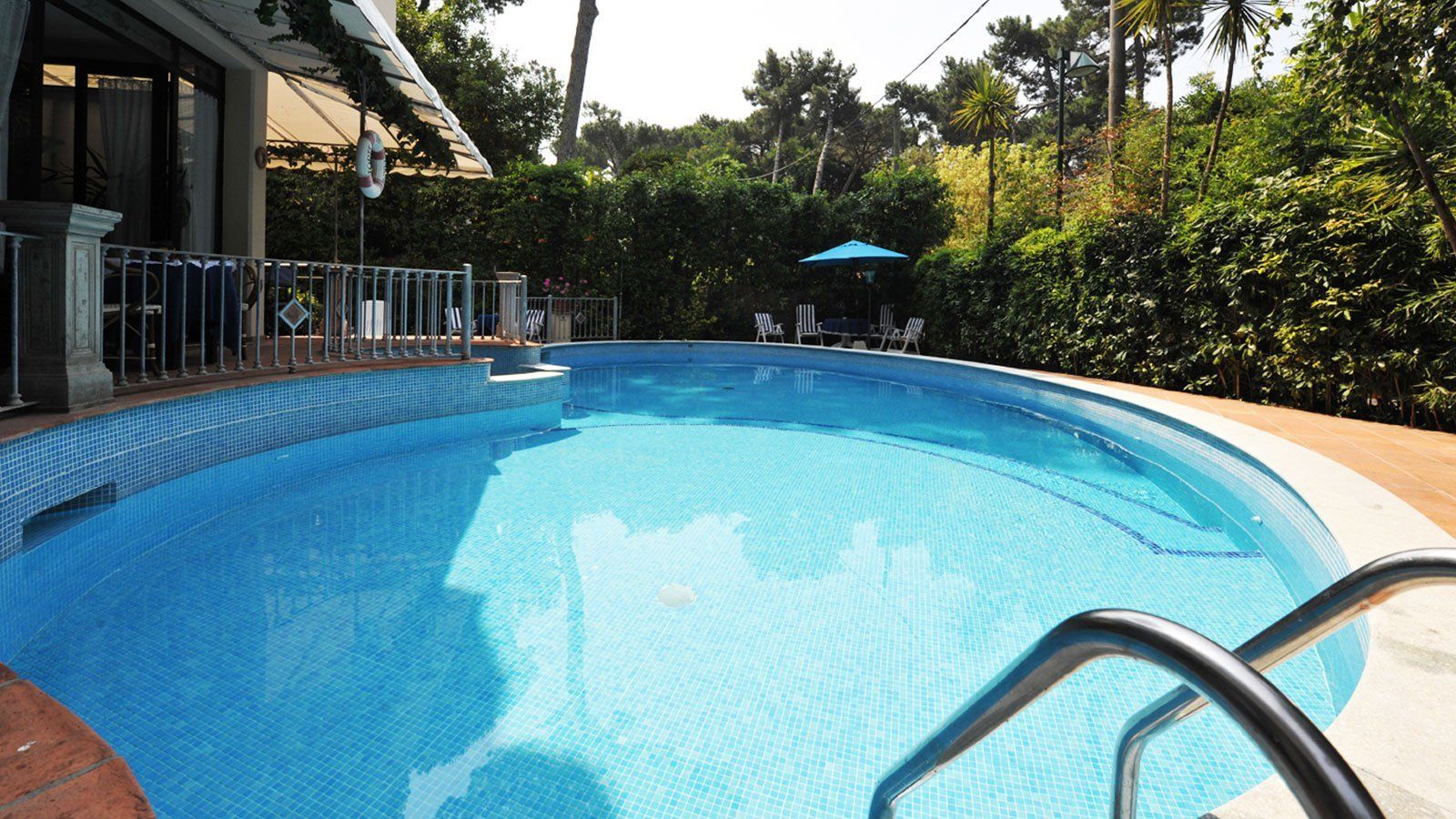 Piscina blu dalle forme sinuose accanto a un patio ombreggiato e a una siepe verde in una giornata di sole.