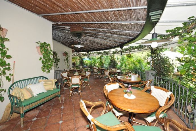 Patio esterno con sedie in vimini, tavolini rotondi in legno e una rigogliosa vegetazione sotto una tettoia ombreggiante.