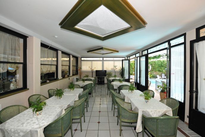 Luminosa sala da pranzo con sedie verdi, tovaglie bianche e grandi finestre che si affacciano su un giardino.