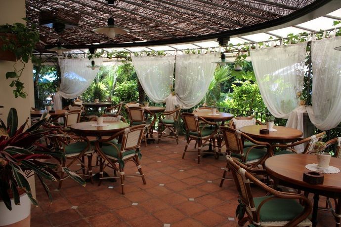 Terrazza all'aperto con tavoli e sedie in legno, tende bianche e una vegetazione rigogliosa.