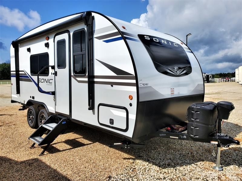 White and gray Sonic travel trailer parked on gravel, with steps down and blue accents.