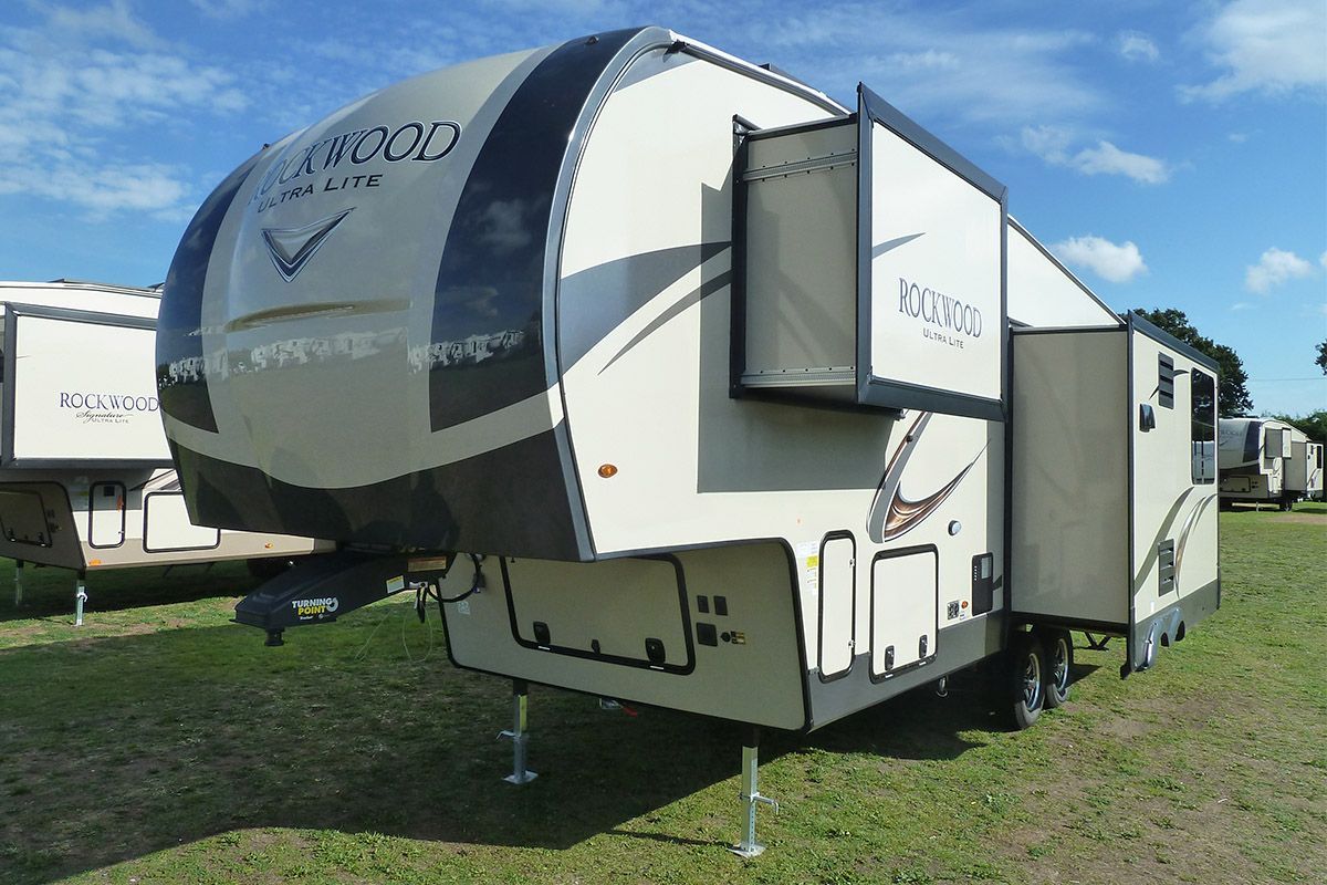 Tan and gray fifth-wheel RV with a slide-out, parked on green grass.