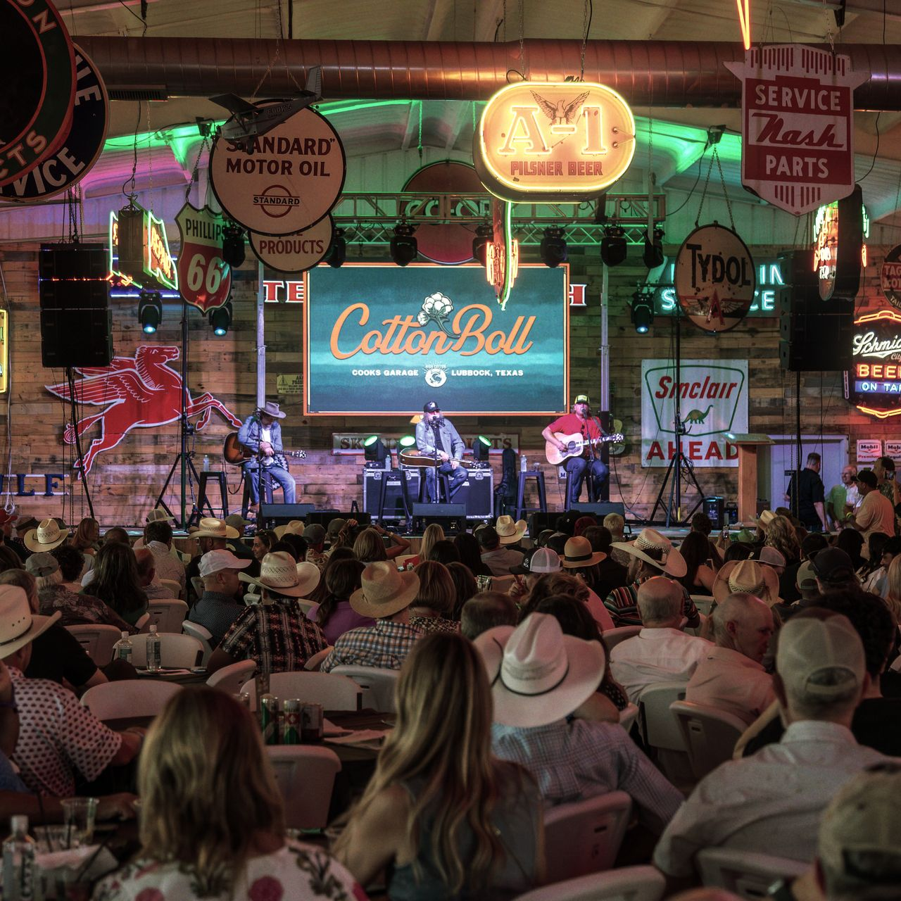 Band performing on stage at a venue called Cotton Boll, decorated with neon signs; audience seated.