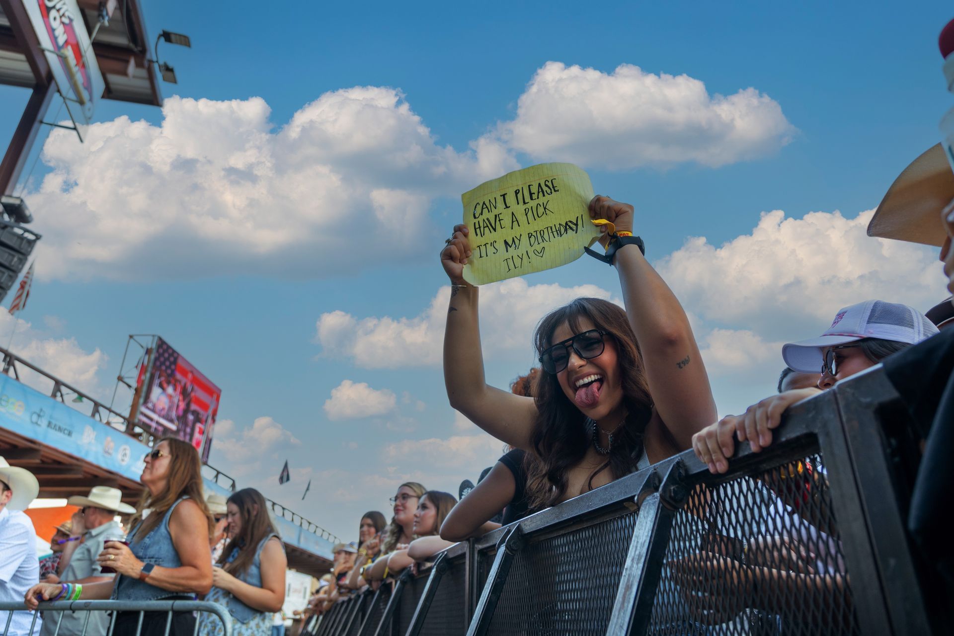 Woman holding a sign at outdoor concert, smiling, sticking out her tongue, blue sky with clouds in background.