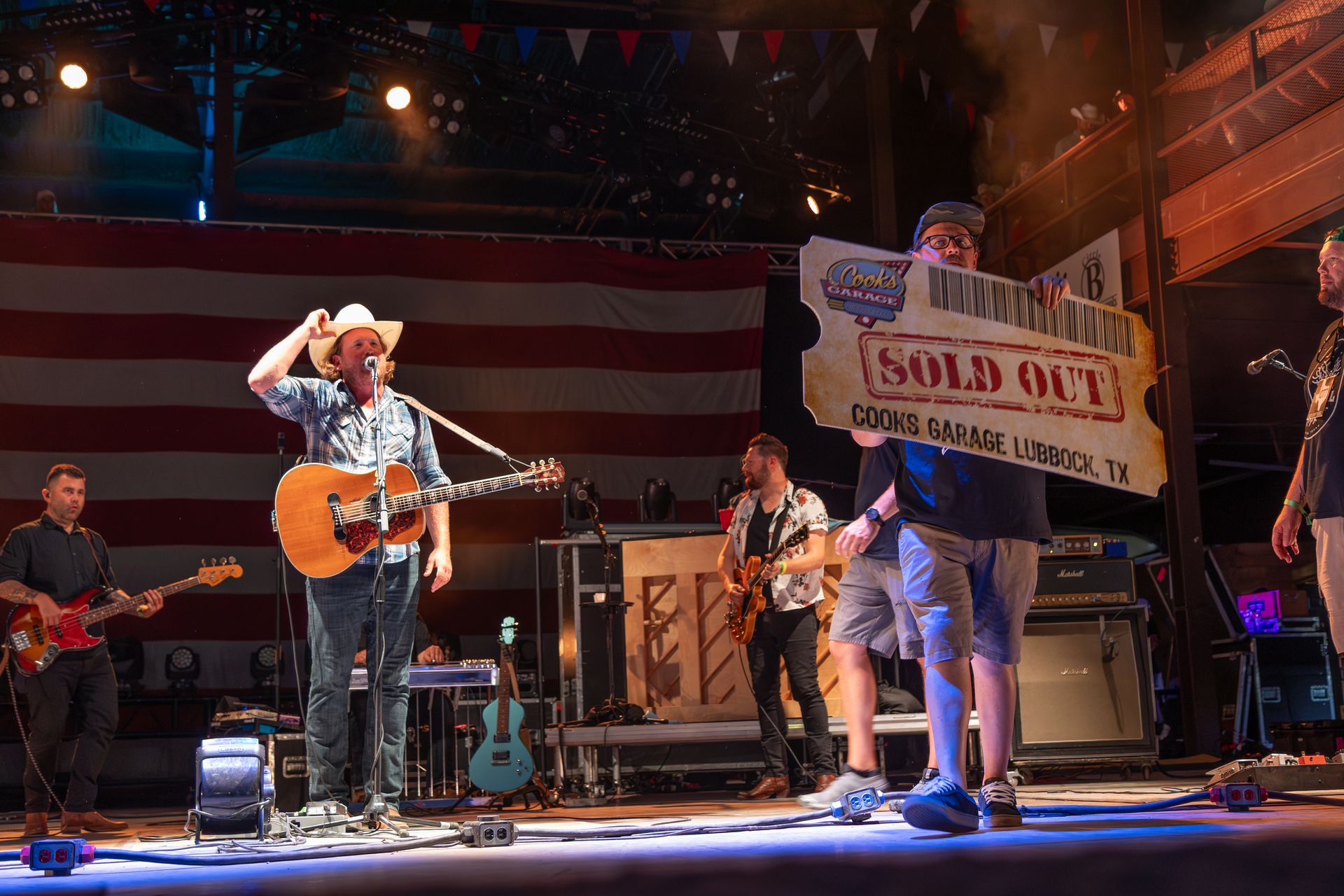 Band performs on stage with "Sold Out" sign at a concert venue, American flag backdrop.