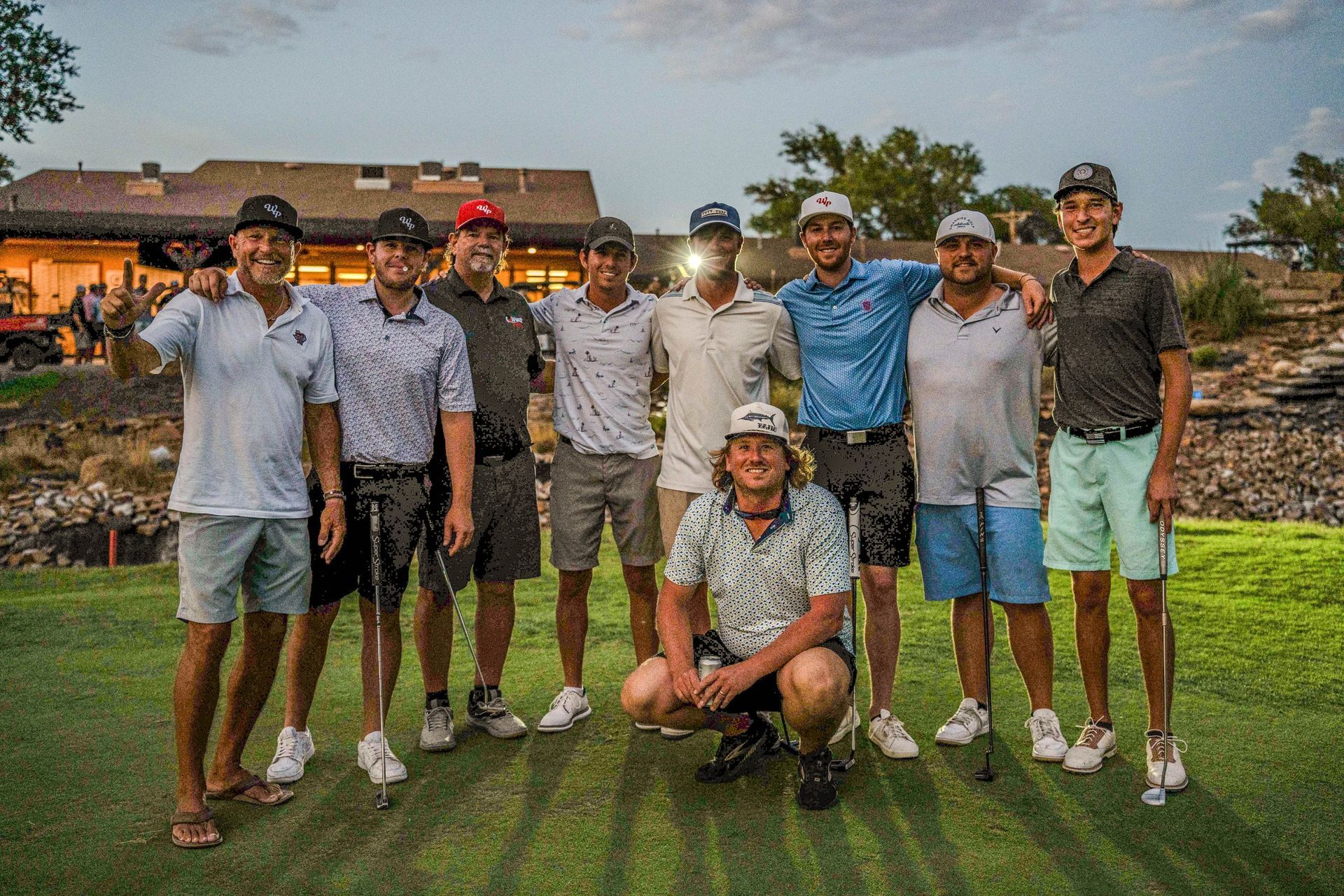 Group of golfers pose on a golf course, smiling with arms around each other after a round.