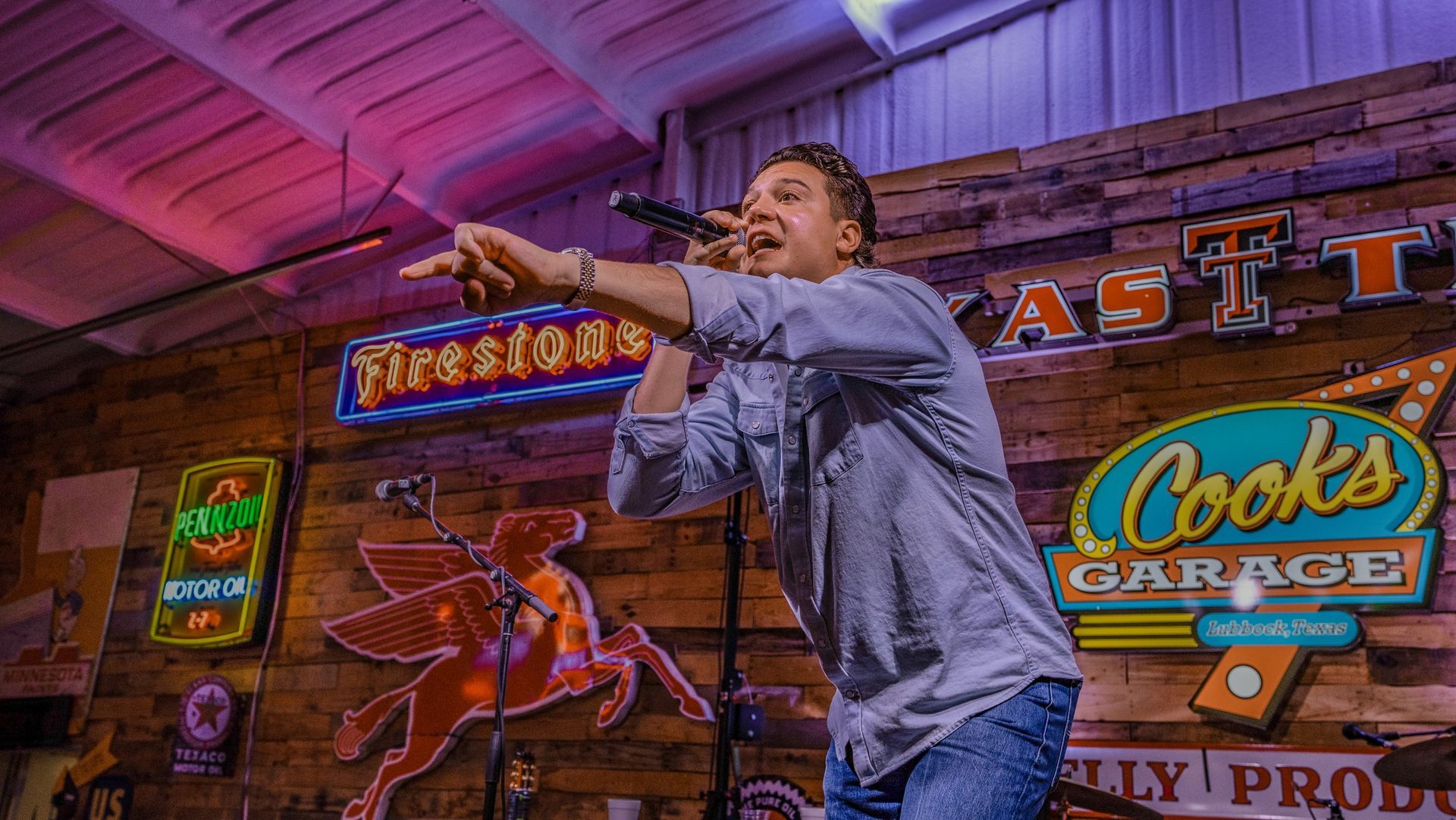 Man singing into a microphone, pointing. He is in a bar with neon signs and a wooden wall.