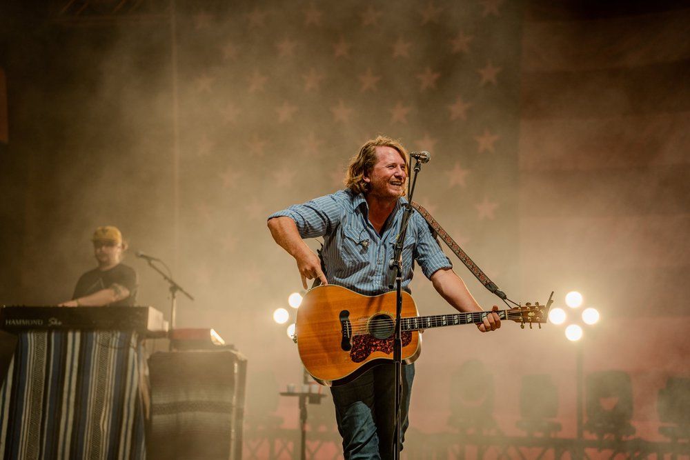 Man playing acoustic guitar on stage, smiling. Stage lights and American flag backdrop.