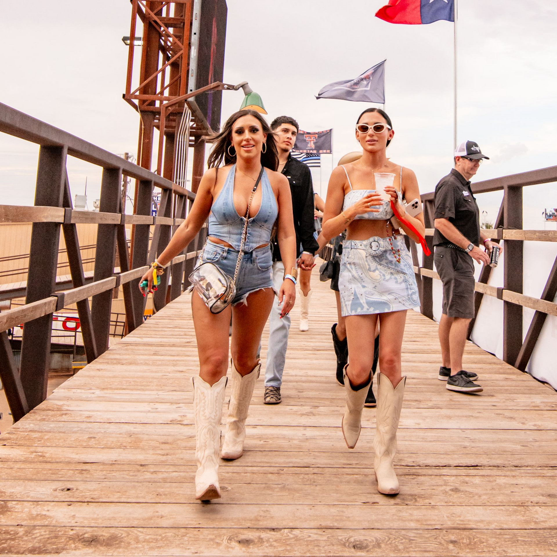 Two women and two men walk on a wooden bridge, outdoors. The women wear denim outfits and cowboy boots.