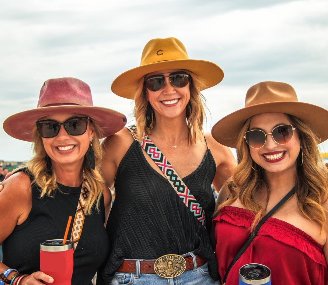 Two women and two men walk on a wooden bridge, outdoors. The women wear denim outfits and cowboy boots.