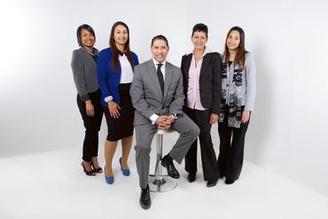 Group of five business professionals posing in front of a white backdrop; man seated, others standing.