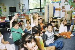 Children in a classroom seated on the floor with hands raised. Bright room with posters and a window.