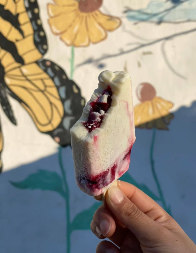 Mano sosteniendo una paleta parcialmente comida, blanca con relleno morado visible, frente a un mural de una mariposa y flores.