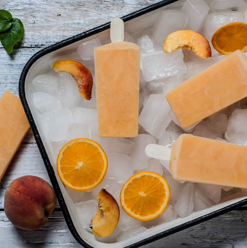 Paletas de melocotón y naranja sobre hielo en una bandeja de metal, con rodajas de fruta.