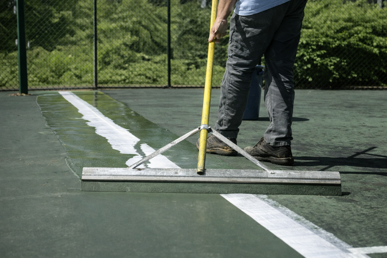 Person smoothing wet coating on a tennis court line with a squeegee. Green court, white line, yellow handle.