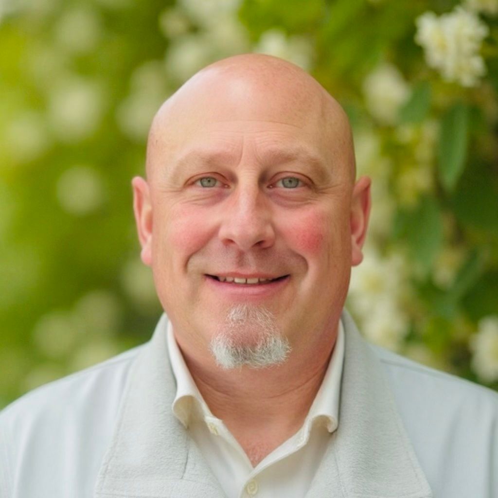 Bald man with goatee smiles in front of green and white foliage. He wears a light gray jacket over a white shirt.