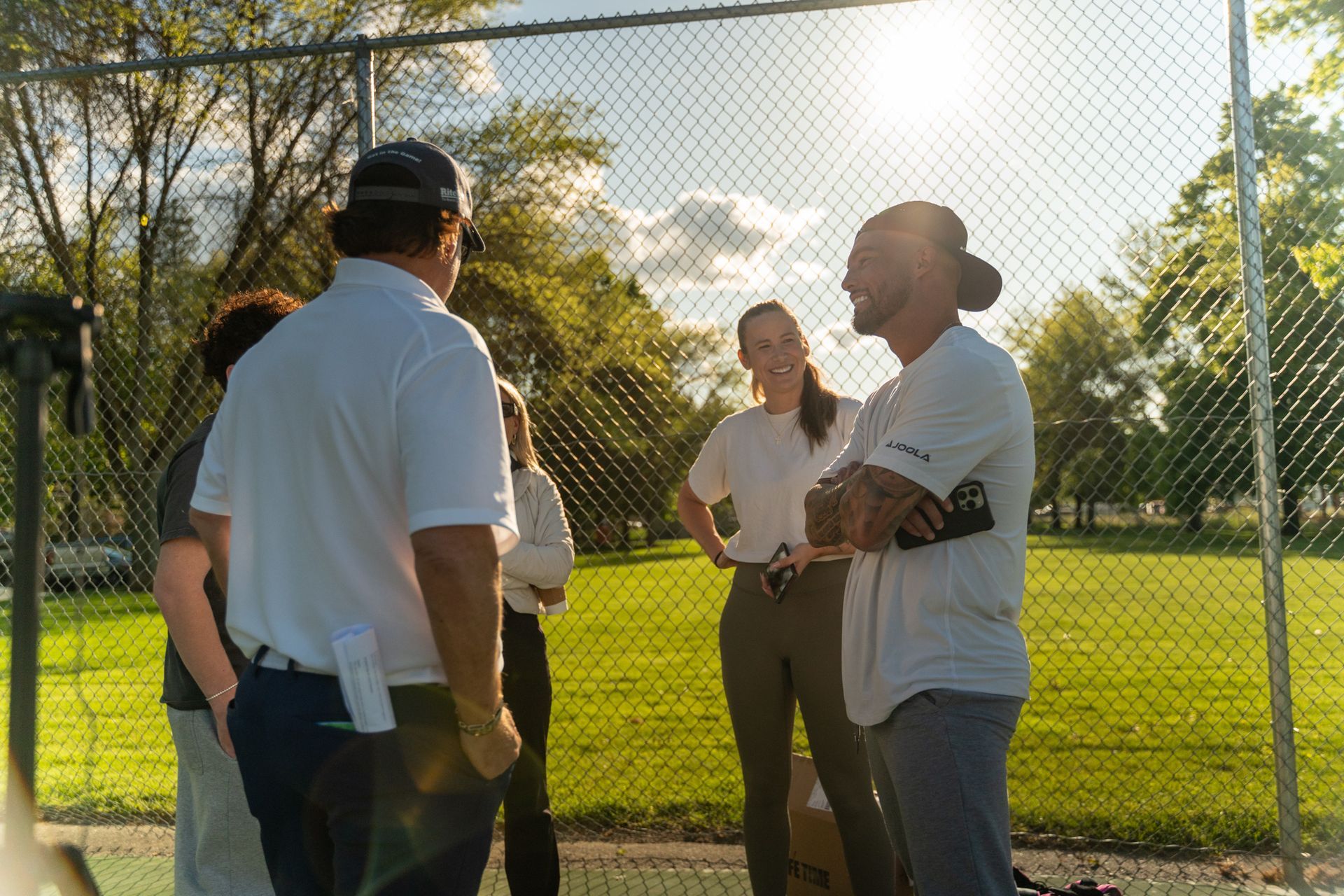 People in white shirts and baseball caps conversing near a chain link fence on a sunny day.