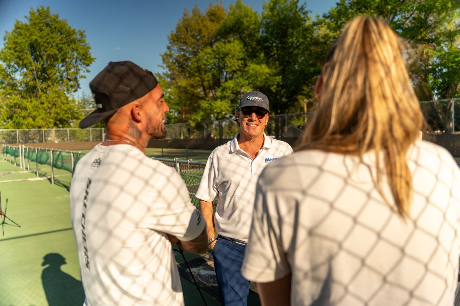Three people converse on a tennis court; one wears a cap and white tee, the other a white polo, and the third has blond hair.