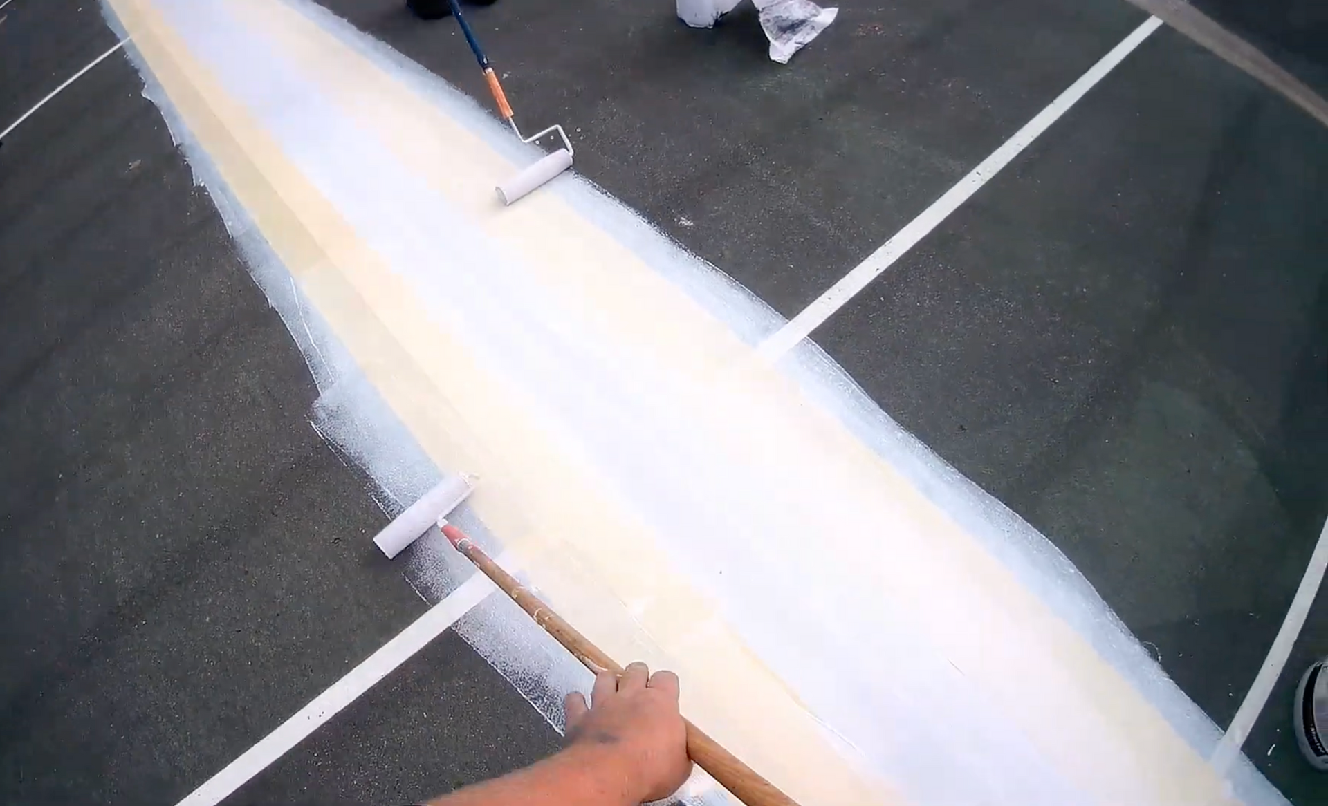 Person smoothing wet coating on a tennis court line with a squeegee. Green court, white line, yellow handle.
