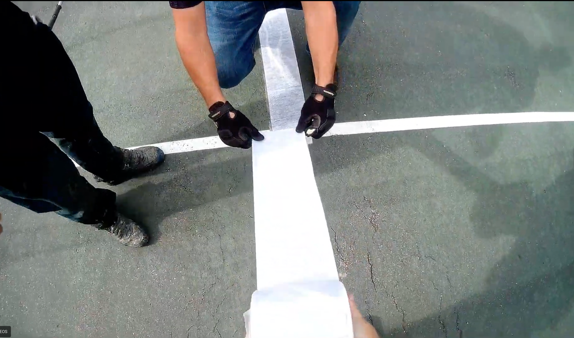 Person kneeling, applying RiteWay tape to a cracked tennis court line, outdoors. Hands in black gloves.