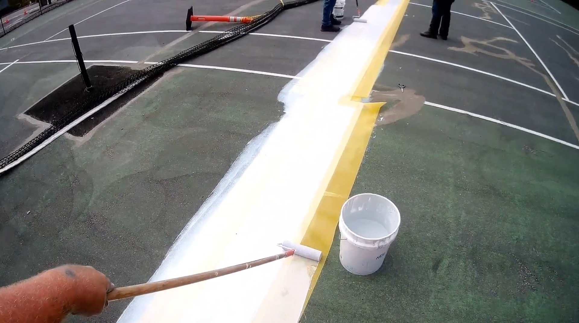 Two men painting white lines on a blue tennis court; one kneels, the other holds a paint roller.