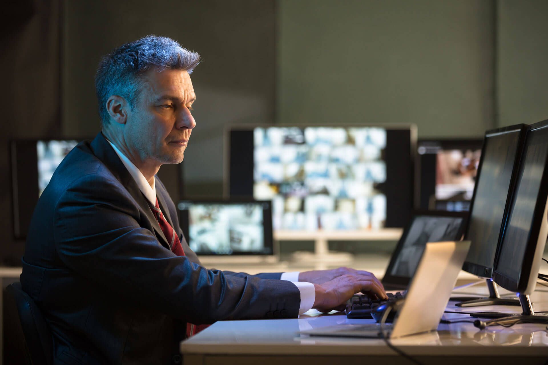 Hombre de traje escribiendo en una computadora con múltiples monitores en una habitación poco iluminada.
