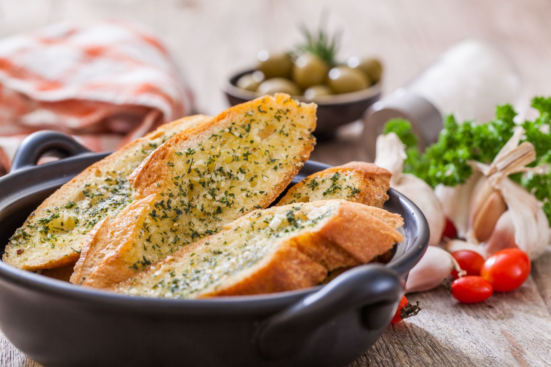 Garlic bread slices in a black bowl, surrounded by olives, garlic cloves, herbs, and tomatoes.