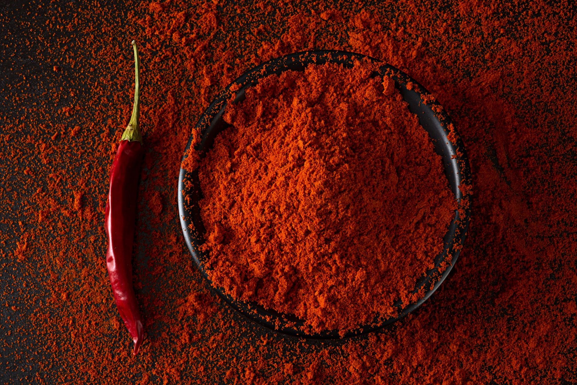 Red chili pepper beside a bowl of red chili powder, on a dark surface.